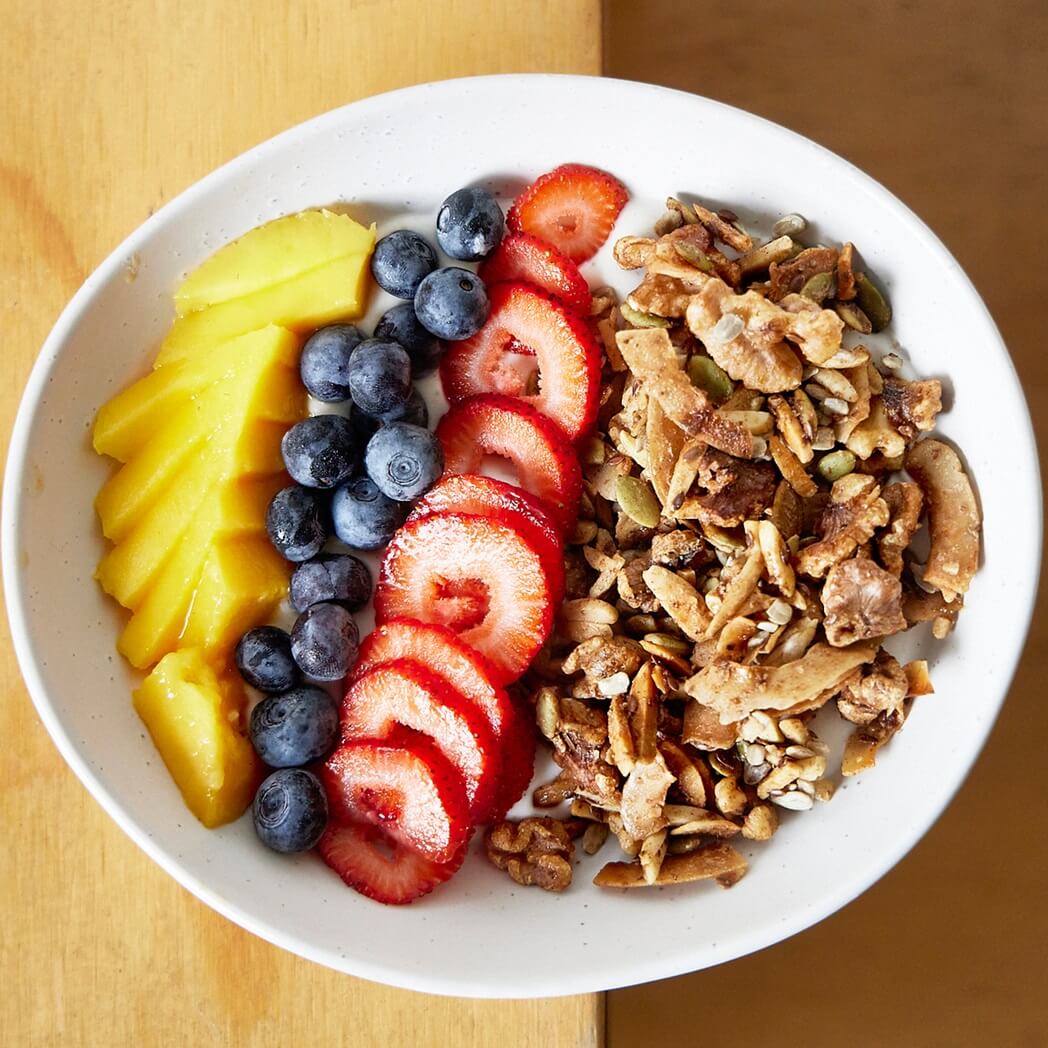 Nutty Granola breakfast with yoghurt, strawberries, blueberries and mango in a white bowl.